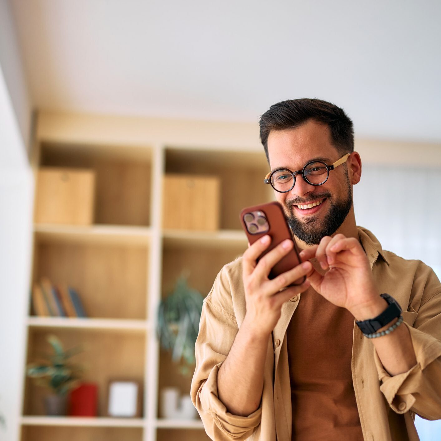 Smiling man using smartphone indoors, wearing glasses and beige jacket.
